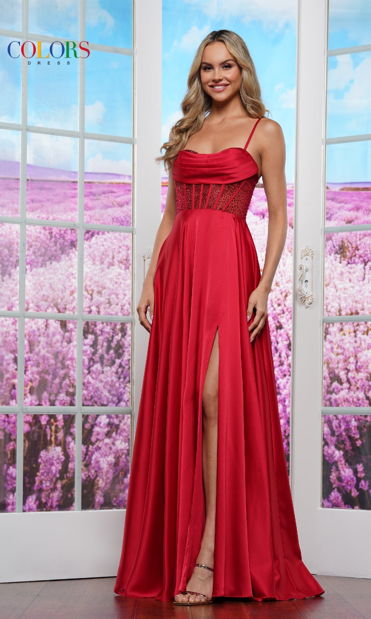 A woman stands indoors before glass doors, wearing the Sheer-Corset Long Prom Dress: Colors Dress 3431, a formal gown with a thigh-high slit; through the doors, a purple flower field is in full bloom.